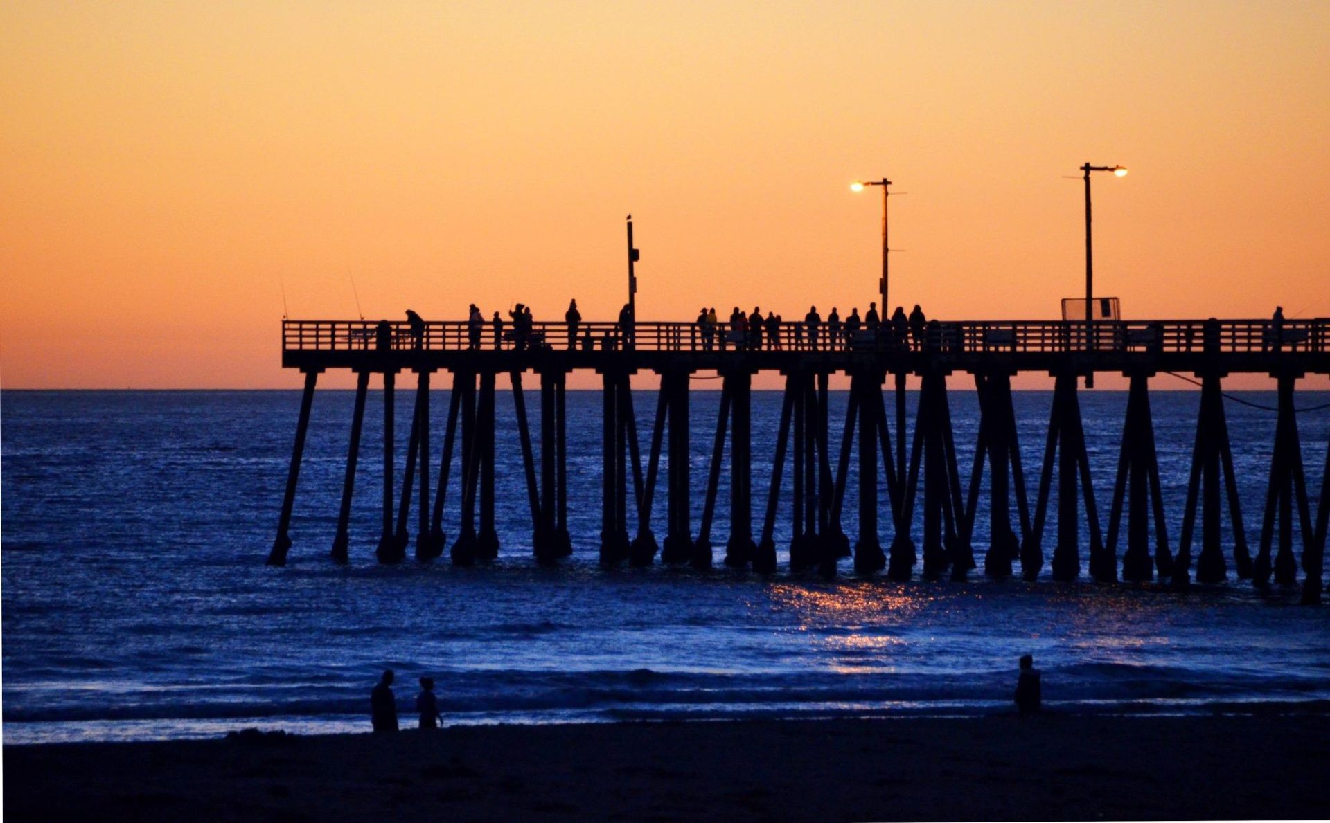A photograph of Pismo Beach Pier at sunset: the pier in black silhouette against an orange sky over dark blue water